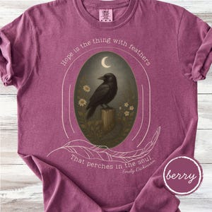 K&ouml;nnte beinhalten: Beerenfarbenes T-Shirt mit einer Grafik, die eine schwarze Kr&auml;he auf einem Holzpfosten zeigt, umgeben von Blumen und einem Halbmond. Der Text lautet: "Hoffnung ist das Ding mit Federn, das in der Seele sitzt."