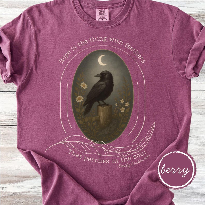 K&ouml;nnte beinhalten: Beerenfarbenes T-Shirt mit einer Grafik, die eine schwarze Kr&auml;he auf einem Holzpfosten zeigt, umgeben von Blumen und einem Halbmond. Der Text lautet: "Hoffnung ist das Ding mit Federn, das in der Seele sitzt."