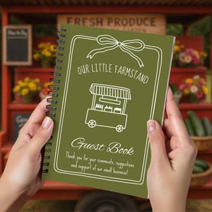 May include: A spiral-bound guest book with a green cover featuring the text "Our Little Farmstand" and a drawing of a farm stand. The words "Guest Book" are below the illustration. The book is held in two hands.