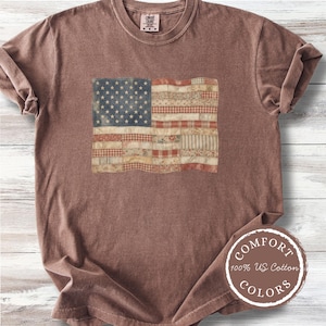 May include: A brown t-shirt featuring a distressed American flag design. The flag is composed of various fabric patterns, with a blue field of stars and red and white stripes. The shirt has a crew neck and short sleeves. The tag says "Comfort Colors 100% US Cotton".