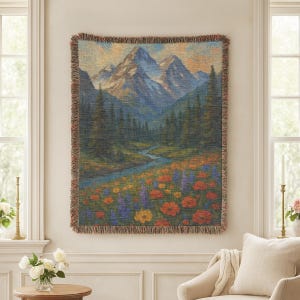 May include: A woven tapestry showing a mountain landscape with a river and wildflowers. The artwork features a mountain range with snow-capped peaks, a winding river, and a field of colourful flowers in shades of orange, yellow, and purple. The tapestry has a fringed edge.