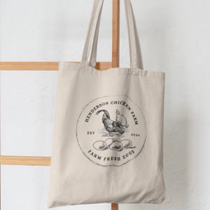 May include: A white canvas tote bag with a black and white logo that says "Henderson Chicken Farm Est. 2024 Farm Fresh Eggs". The logo features a rooster and hens with eggs.
