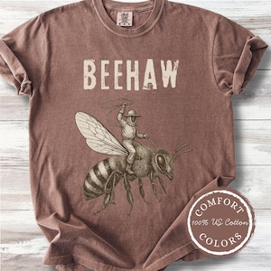 May include: A brown short-sleeve t-shirt featuring the text BEEHAW in a cream-colored font. Below the text is a detailed illustration of a person in a beekeeping suit riding a large bee while holding a lasso. The shirt has a crew neckline.