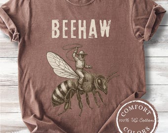 May include: A brown short-sleeve t-shirt featuring the text BEEHAW in a cream-colored font. Below the text is a detailed illustration of a person in a beekeeping suit riding a large bee while holding a lasso. The shirt has a crew neckline.