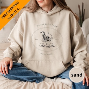 May include: A beige hoodie with a black and white illustration of a rooster and eggs. The hoodie has the text "Henderson Farm" and "Fresh Eggs" printed on it.
