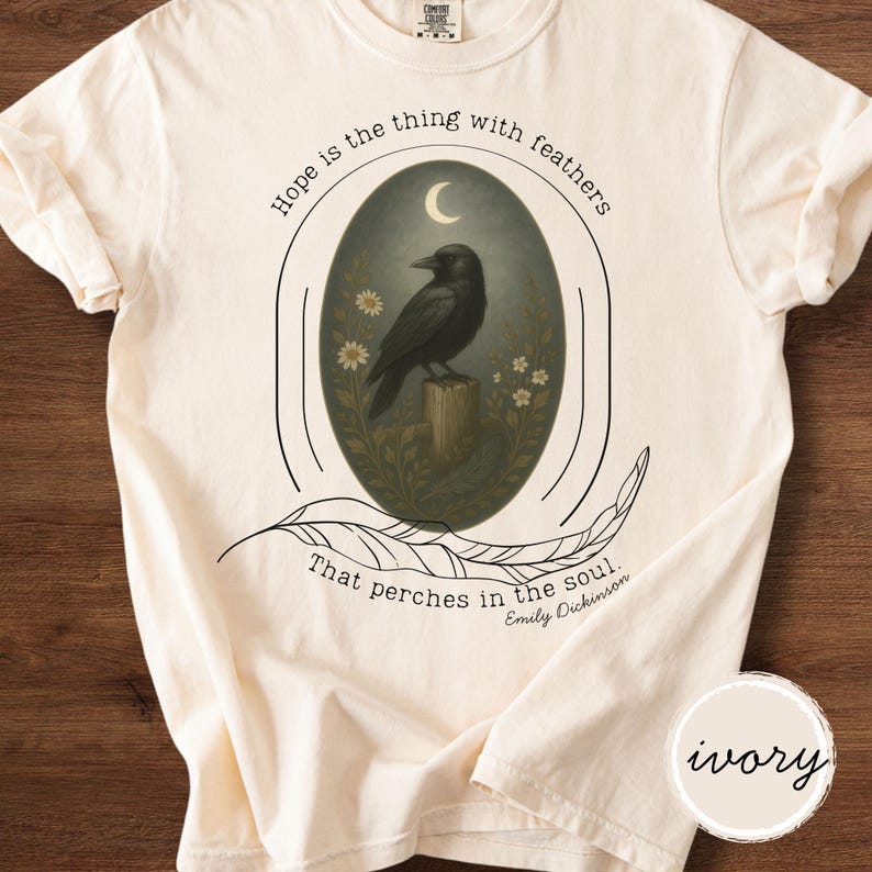 K&ouml;nnte beinhalten: Elfenbeinfarbenes T-Shirt mit einem schwarzen Raben-Design in einem ovalen Rahmen. Das Design beinhaltet einen Mond, Blumen und eine Feder, mit dem Zitat "Hope is the thing with feathers" und "That perches in the soul. Emily Dickinson."