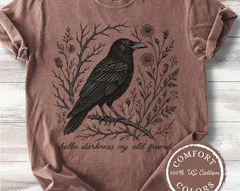Pode incluir: Camiseta marrom com um desenho de corvo preto empoleirado em um galho, cercado por ilustrações florais e de galhos. O texto "hello darkness my old friend" está impresso abaixo. Etiqueta Comfort Colors.
