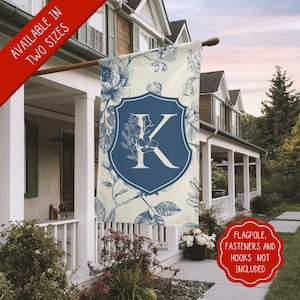 Puede incluir: Una bandera de jardín de color crema con un escudo azul que presenta la letra K en un diseño floral. La bandera está decorada con patrones florales azules y se muestra en un porche blanco.