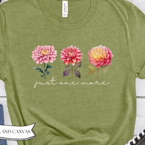 May include: Green t-shirt with a floral design featuring three dahlias in pink, red, and yellow hues. The phrase "just one more" is written in cursive script below the flowers. The shirt is made by Bella and Canvas.