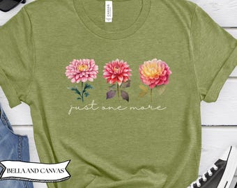 May include: Green t-shirt with a floral design featuring three dahlias in pink, red, and yellow hues. The phrase "just one more" is written in cursive script below the flowers. The shirt is made by Bella and Canvas.