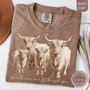 May include: A brown Comfort Colors t-shirt featuring a graphic of four Highland cows. The shirt has the text "Live like someone left the gate open." A silver necklace and a Comfort Colors logo are also visible.
