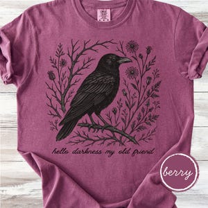 May include: Berry-colored t-shirt featuring a detailed black illustration of a crow perched on a branch, surrounded by floral and branch elements. The text "hello darkness my old friend" is printed below the crow. The shirt has a crew neck and short sleeves.