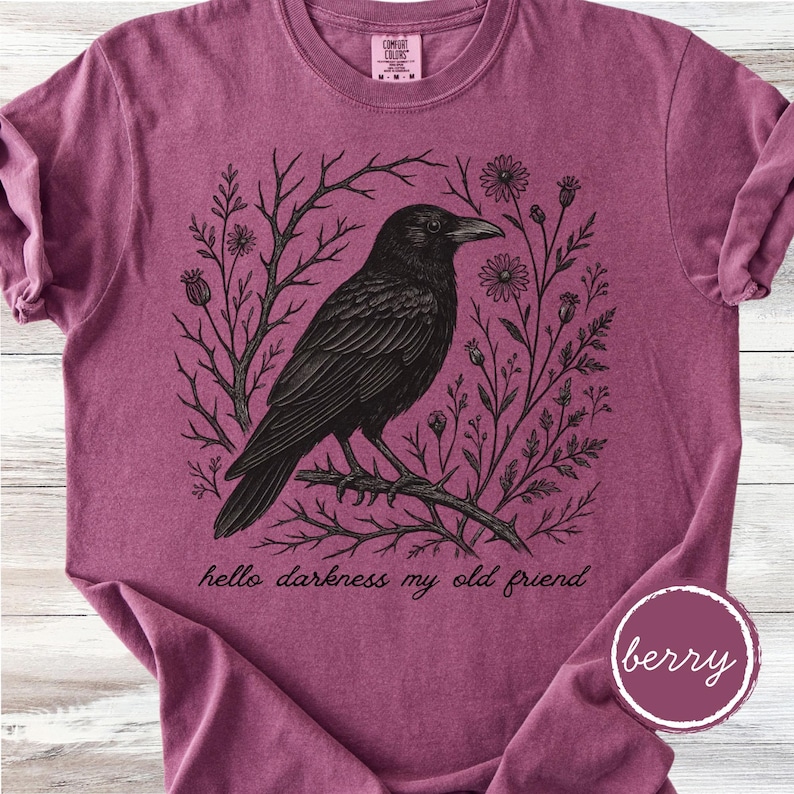 May include: Berry-colored t-shirt featuring a detailed black illustration of a crow perched on a branch, surrounded by floral and branch elements. The text "hello darkness my old friend" is printed below the crow. The shirt has a crew neck and short sleeves.
