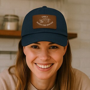 Peut inclure: Une casquette de baseball bleu marine avec un patch en cuir marron qui dit "Emily Makes the Dough" avec une petite illustration d'une miche de pain et de fleurs.