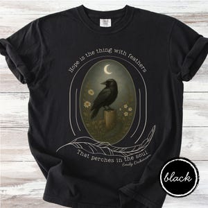 K&ouml;nnte beinhalten: Schwarzes T-Shirt mit einem ovalen Design, das eine schwarze Kr&auml;he auf einem Baumstumpf unter einem Halbmond zeigt. Das Design enth&auml;lt das Zitat "Hoffnung ist das Ding mit Federn, das in der Seele haust" von Emily Dickinson.
