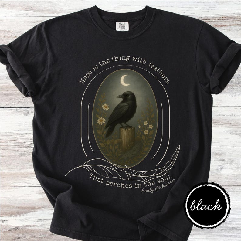 K&ouml;nnte beinhalten: Schwarzes T-Shirt mit einem ovalen Design, das eine schwarze Kr&auml;he auf einem Baumstumpf unter einem Halbmond zeigt. Das Design enth&auml;lt das Zitat "Hoffnung ist das Ding mit Federn, das in der Seele haust" von Emily Dickinson.