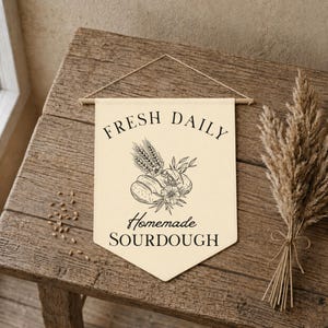 May include: Cream-coloured banner with the words "FRESH DAILY Homemade SOURDOUGH" in black. The banner features a black illustration of bread, wheat, and flowers. The banner is suspended by a wooden dowel and twine.