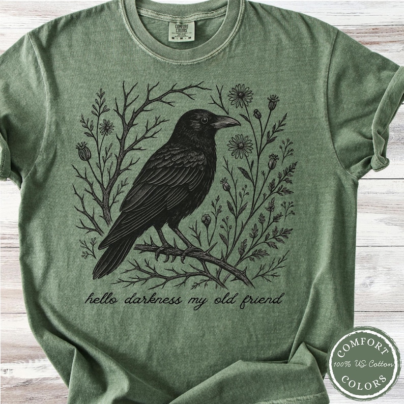 May include: Green t-shirt featuring a detailed black raven perched on a branch, surrounded by floral and botanical elements. The text "hello darkness my old friend" is printed below the raven. The shirt has a Comfort Colors label.