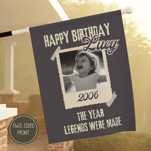 Könnte beinhalten: Eine zweiseitige, dunkelgraue Flagge mit dem weißen Text "HAPPY BIRTHDAY Lucy". In der Mitte befindet sich ein Schwarz-Weiß-Foto eines Kindes, darunter das Jahr "2006". Der Text "THE YEAR LEGENDS WERE MADE" steht unten.