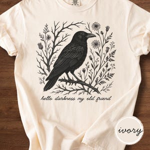 May include: Ivory-colored t-shirt featuring a black illustration of a crow perched on a branch, surrounded by floral and botanical elements. The text "hello darkness my old friend" is printed below the crow. The shirt has short sleeves.