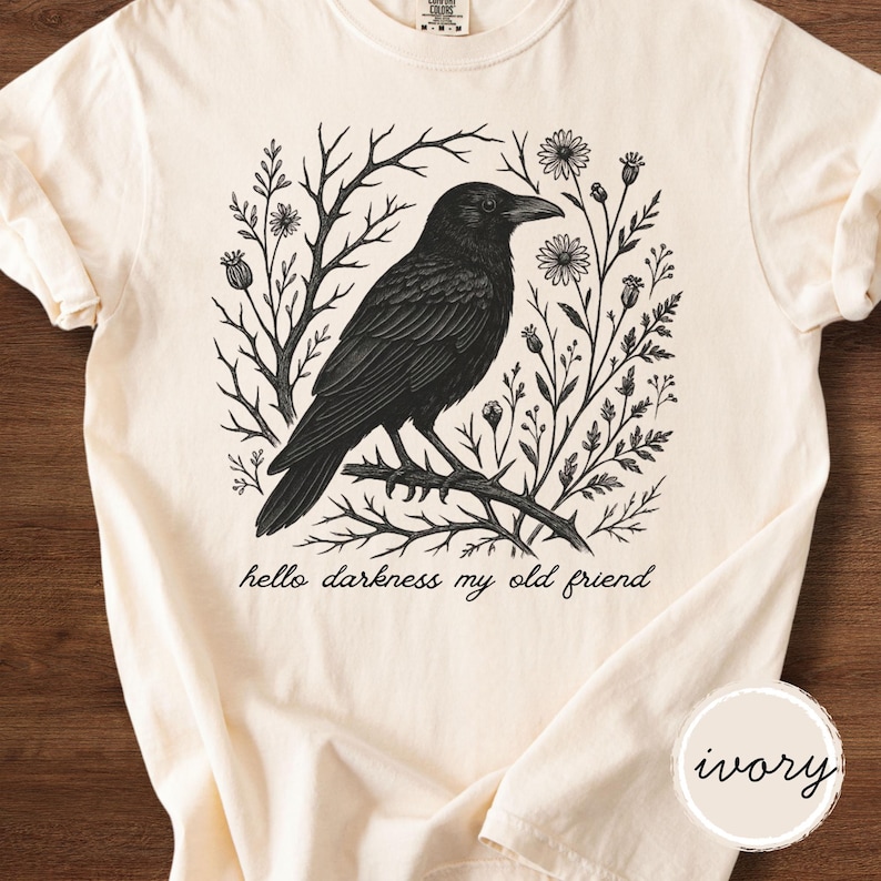 May include: Ivory-colored t-shirt featuring a black illustration of a crow perched on a branch, surrounded by floral and botanical elements. The text "hello darkness my old friend" is printed below the crow. The shirt has short sleeves.