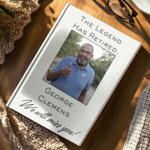 Puede incluir: Un libro blanco con el texto "THE LEGEND HAS RETIRED." y "GEORGE CLEMENS We will miss you!" Una foto de un hombre haciendo una señal de pulgar hacia arriba está en el centro. Gafas negras sobre la superficie de madera.