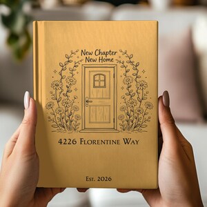 May include: A mustard-colored journal with a floral door design. The text reads "New Chapter New Home" above the door, with "4226 Florentine Way" and "Est. 2026" below. The journal is held by two hands.