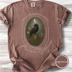 K&ouml;nnte beinhalten: Braunes T-Shirt mit einem zentralen ovalen Design. Es zeigt eine schwarze Kr&auml;he auf einem Holzpfosten unter einem Halbmond, mit dem Zitat "Hoffnung ist das Ding mit Federn, das in der Seele sitzt" von Emily Dickinson.