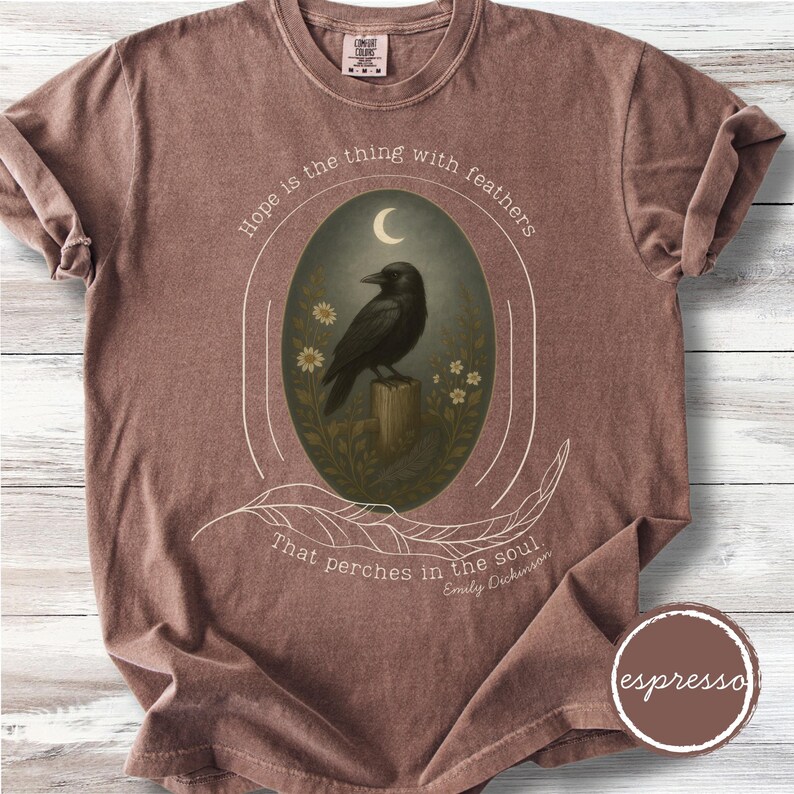 K&ouml;nnte beinhalten: Braunes T-Shirt mit einem zentralen ovalen Design. Es zeigt eine schwarze Kr&auml;he auf einem Holzpfosten unter einem Halbmond, mit dem Zitat "Hoffnung ist das Ding mit Federn, das in der Seele sitzt" von Emily Dickinson.