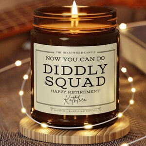 May include: A lit brown glass candle jar with a white label that reads "Now You Can Do Diddly Squad Happy Retirement Kathleen". The candle sits on a wooden coaster, surrounded by string lights. The candle is made of vegan, ecofriendly, natural soy.