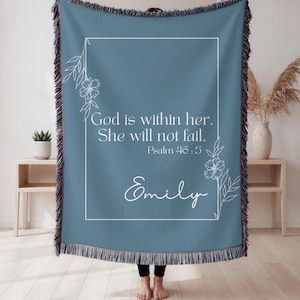 Puede incluir: Una manta tejida azul con un diseño floral blanco y el texto "God is within her. She will not fail. Psalm 46:5 Emily." La manta tiene un borde con flecos y se muestra en interiores.