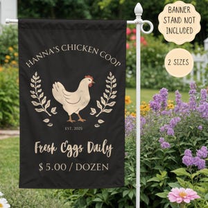 May include: Black garden flag with white text that reads "Hanna's Chicken Coop Est. 2025 Fresh Eggs Daily $5.00/DOZEN". A white chicken illustration with red comb and wattle is centered on the flag. The chicken is surrounded by two green leafy branches.