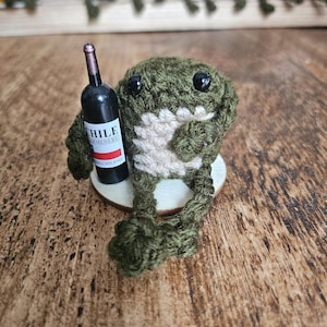 May include: A small, dark green crocheted frog figurine with a cream-coloured belly and black bead eyes. The frog is holding a miniature black wine bottle with a label that reads CHILE CARMENERE VINO CHILENO, sitting on a circular wooden base.
