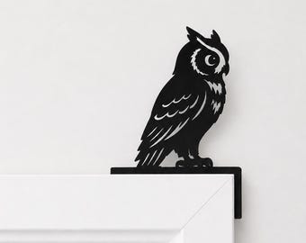 Puede incluir: Una silueta de búho de metal negro diseñada para colocarse en la esquina de un marco de puerta. El búho se muestra de perfil con detalles de plumas recortadas y un ojo circular, con una base en forma de L que se apoya sobre una superficie.