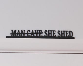 Puede incluir: Letrero de metal negro con las palabras "MAN CAVE SHE SHED" sobre una puerta blanca. El letrero está montado en el marco de la puerta. El fondo es una pared blanca lisa.
