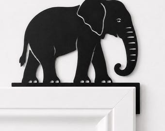 Puede incluir: Una silueta de elefante de metal negro diseñada para descansar en la esquina superior del marco de una puerta. El elefante se muestra de perfil, mirando hacia la derecha, con la trompa curvada hacia abajo y las orejas y patas claramente definidas.