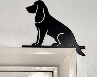 Puede incluir: Una silueta negra de un perro en posición sentada, diseñada para montarse en la esquina del marco de una puerta. El recorte presenta el perfil distintivo de un sabueso con orejas largas, cuello delgado y cola a lo largo de la base.