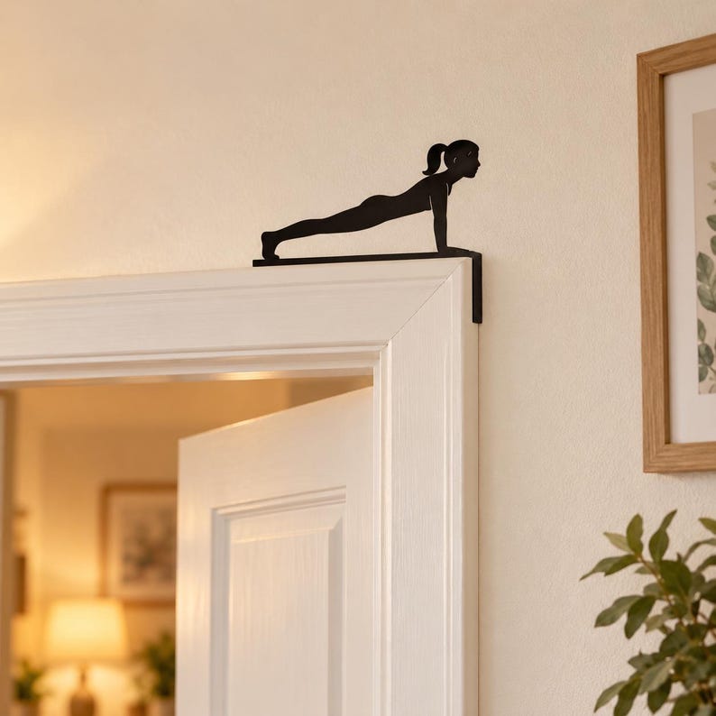 K&ouml;nnte beinhalten: Ein T&uuml;rschmuck aus schwarzem Metall, der die Silhouette einer Person in der Plank-Position zeigt. Das flache, L-f&ouml;rmige Dekorationselement ist so gestaltet, dass es sicher auf der oberen Ecke eines T&uuml;rrahmens aufliegt.