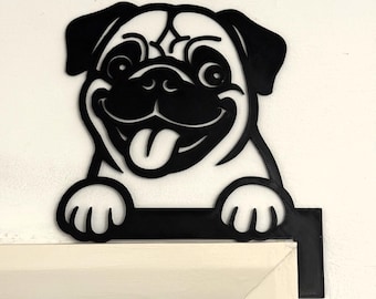 Puede incluir: Decoración de perro pug de metal negro con una cara sonriente y la lengua fuera. El pug se aferra a un marco de puerta blanco. Los rasgos del perro son detallados y el diseño está recortado.