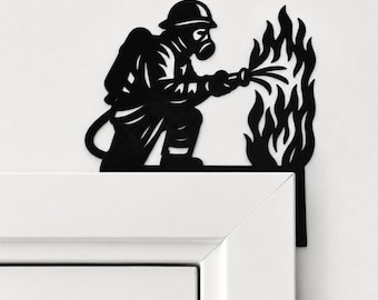 Puede incluir: Una pieza de arte mural de metal negro con la forma de un bombero con casco y máscara de gas, arrodillado mientras sostiene una manguera para rociar agua sobre una llama estilizada. La silueta está diseñada para el marco de una puerta.