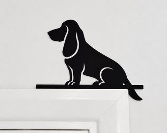 Puede incluir: Una silueta de metal negro de un perro basset hound sentado con orejas largas y cola, diseñada para montarse en la esquina superior del marco de una puerta. La figura plana y negra presenta una barra base horizontal sobre el marco.