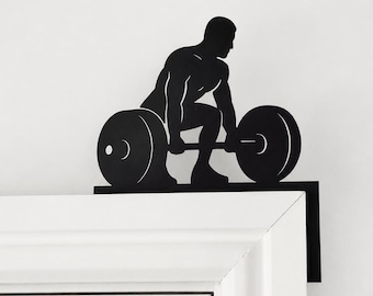 Könnte beinhalten: Ein Tür-Aufsatz aus schwarzem Metall mit der Silhouette einer Person, die ein Kreuzheben mit einer Langhantel ausführt. Das flache, abgewinkelte Design ist so geformt, dass es fest auf die obere Ecke eines Türrahmens passt, in Schwarz.
