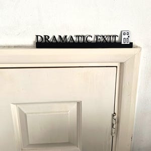 May include: A black sign that reads "DRAMATIC EXIT" in a stylized font, with a small door icon on the right. The sign is mounted above a white door. The door has a rectangular panel and a visible hinge.