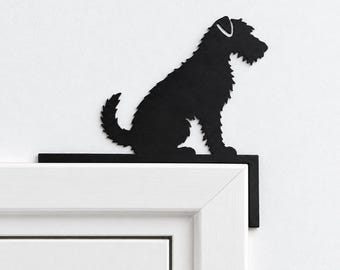 Puede incluir: Un adorno de puerta de metal negro con la silueta de un perro terrier sentado. La pieza plana y angular está diseñada para descansar sobre la esquina superior del marco de una puerta, mostrando el contorno detallado del pelaje.