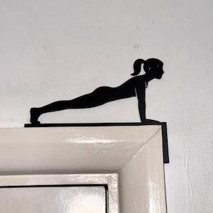 Puede incluir: Silueta metálica negra de una persona en posición de plancha, montada en un marco de puerta blanco. La figura está en una postura de yoga o fitness, con los brazos extendidos y el cuerpo recto. El fondo es una pared blanca texturizada.