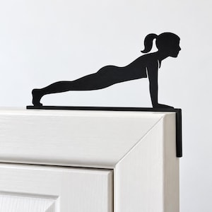 K&ouml;nnte beinhalten: Ein schwarzer T&uuml;raufsatz aus Metall mit der Silhouette einer Person in Plank-Position. Das flache, ausgeschnittene Design ist auf der Oberkante eines T&uuml;rrahmens montiert, wobei ein Winkelst&uuml;ck an der Seite der T&uuml;rzarge nach unten verl&auml;uft.