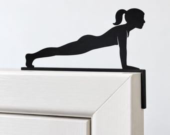 Puede incluir: Un adorno de puerta de metal negro con la silueta de una persona en posición de plancha. El diseño plano y recortado está montado en el borde superior del marco de una puerta, con un soporte en ángulo recto que baja por el lateral.