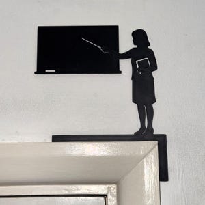 May include: Black metal silhouette of a teacher with a pointer, standing near a chalkboard. The teacher is holding a book. The decorative piece is designed to be placed on a door frame.
