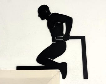 Puede incluir: Silueta negra de metal de una persona haciendo un ejercicio de flexiones en una barra. La figura está montada en una pared blanca, con la barra que se extiende desde la pared. La obra de arte representa a una persona en una pose de entrenamiento, lo que sugiere un tema de fitness o gimnasio.
