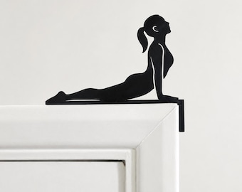 Könnte beinhalten: Eine schwarze Metallsilhouette einer Person in der Kobra-Yoga-Pose, die so gestaltet ist, dass sie auf der Oberkante eines Türrahmens aufliegt. Die Figur hat einen Pferdeschwanz und ist horizontal mit nach oben gestreckten Armen positioniert.